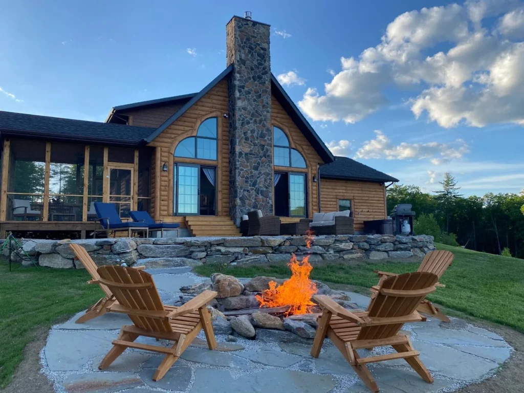 Adirondack chairs around a blazing stone fire pit outside a two-story log home with a large stone chimney