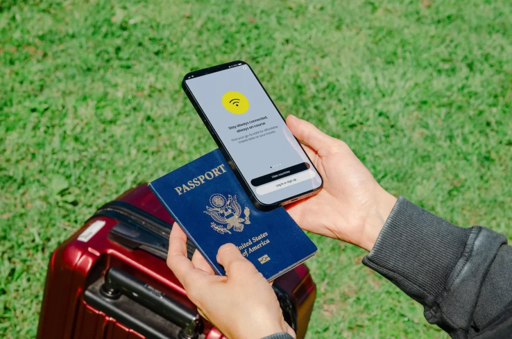 Hands holding a US passport and a smartphone showing a travel eSIM app next to a suitcase
