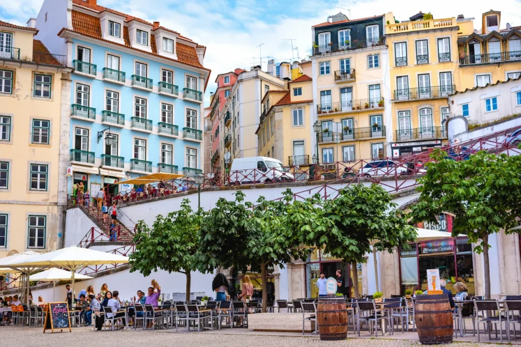 A bustling plaza square with outdoor dining tables below colorful, historic apartment buildings in Lisbon