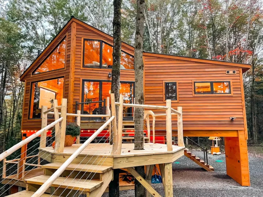 Modern two-story wooden treehouse cabin with large glowing windows surrounded by trees in Vermont