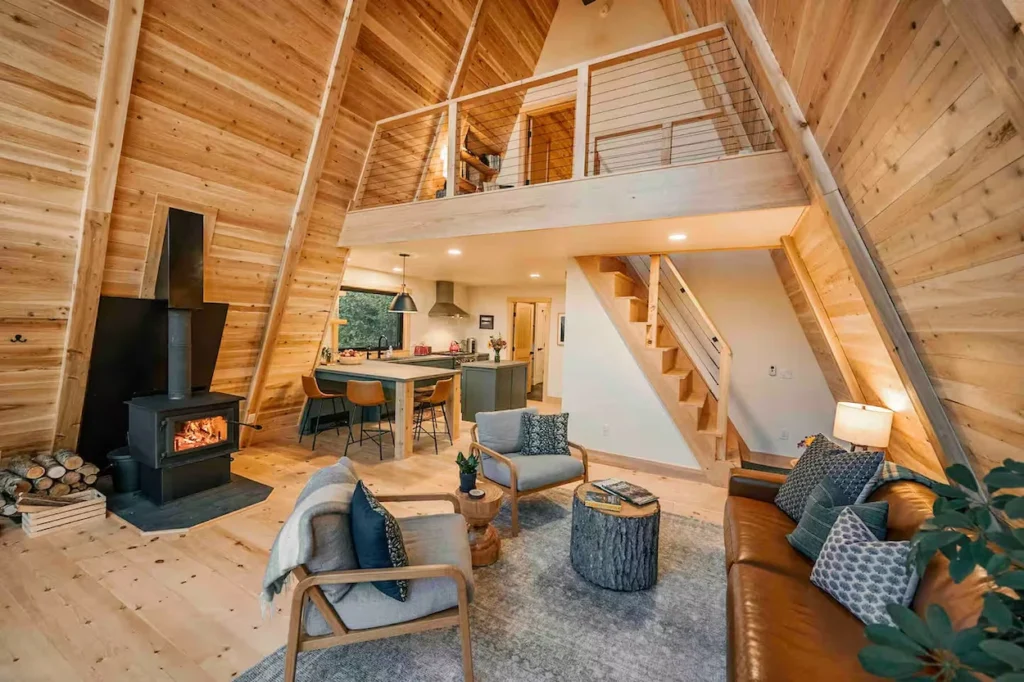 Interior of an A-frame cabin living room featuring light wood paneling, a wood-burning stove, and a loft bedroom overview