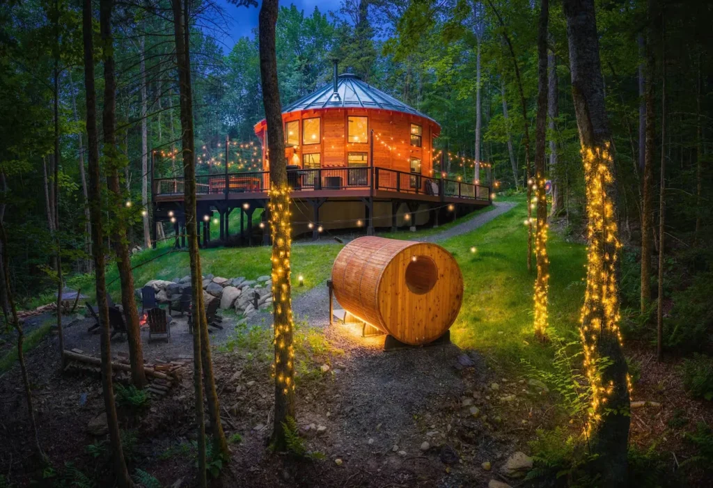 Illuminated wooden yurt and barrel sauna on a hillside at dusk