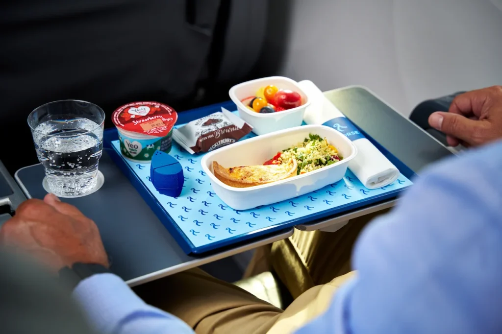 An airplane tray table holds a meal set featuring a main dish with couscous