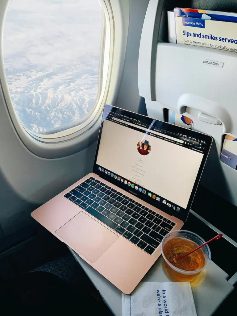 Open laptop on an airplane tray table next to an iced drink by a window looking out at snowy mountains