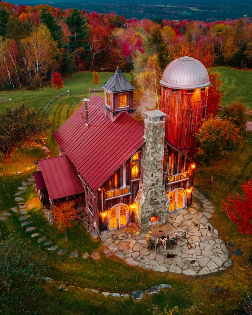 Aerial view of a rustic barn and silo cabin with a stone patio fireplace surrounded by autumn foliage in Vermont
