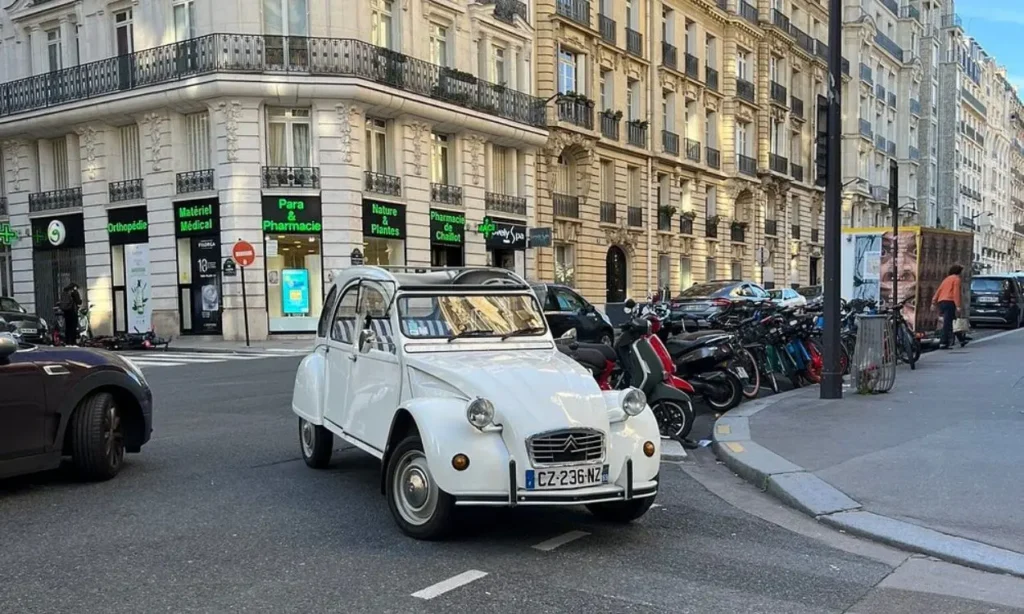 A vintage white Citroën 2CV car driving on a street corner in Paris.