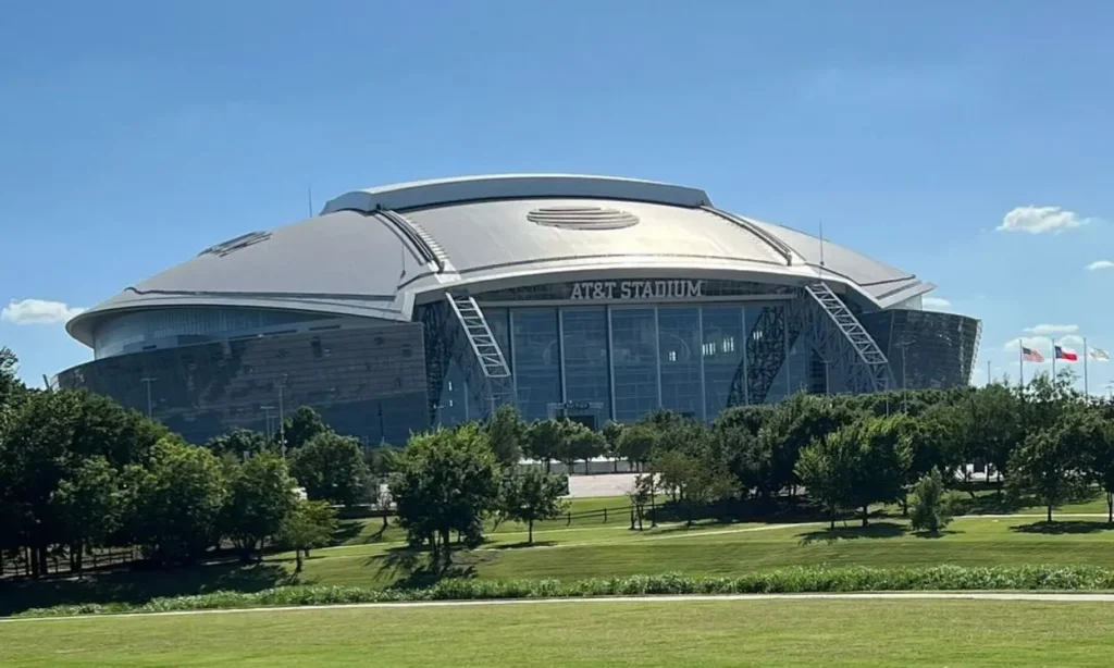 The massive domed exterior of AT&T Stadium with trees in the foreground.