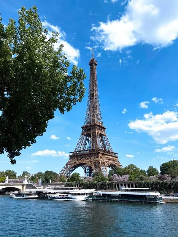 The Eiffel Tower rising above the Seine River on a bright day with boats below.