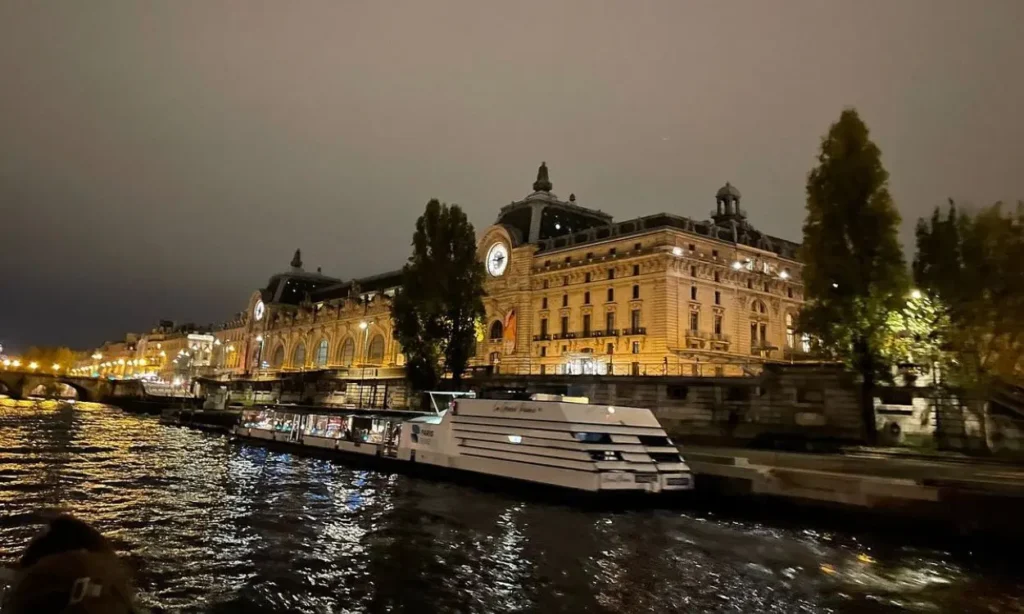 A night view of the illuminated Musée d’Orsay building along the Seine River with boats docked nearby.