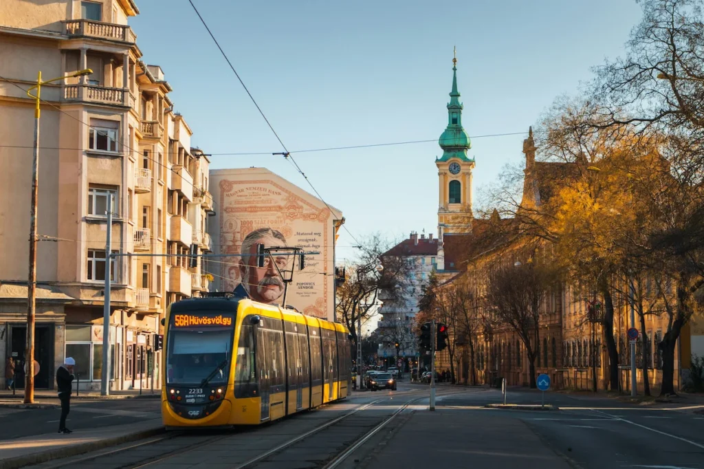Budapest yellow CAF tram passing a large portrait street mural and Saint Catherine of Alexandria Church tower