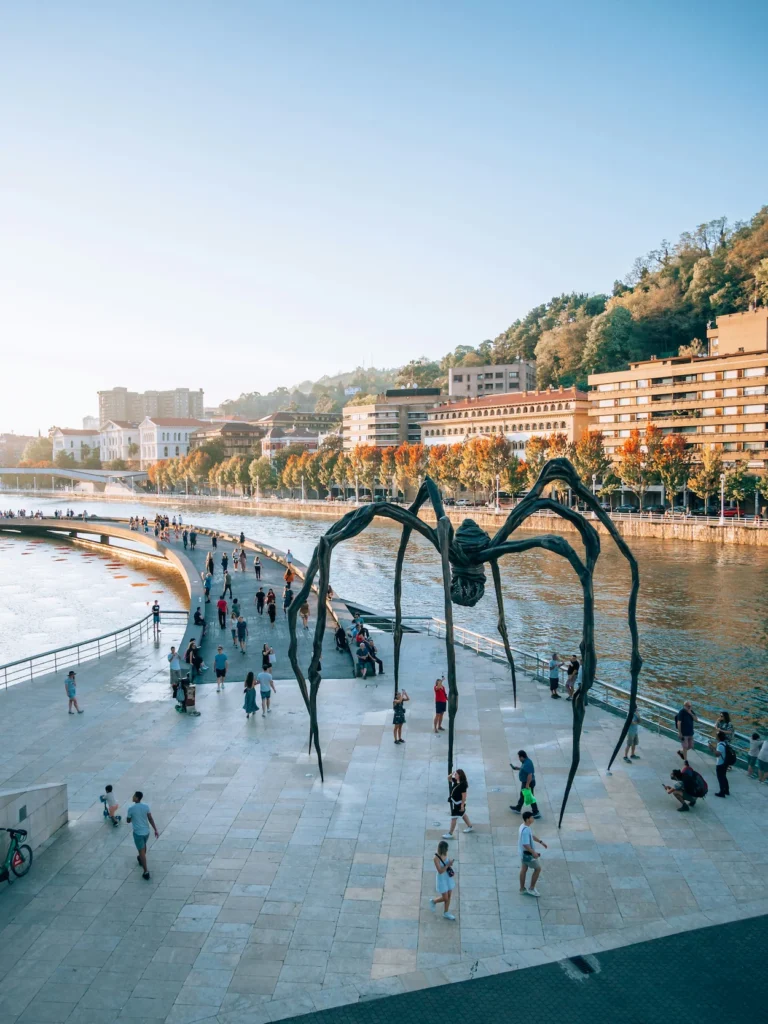 Large bronze spider sculpture standing on a riverside promenade next to a pedestrian walkway