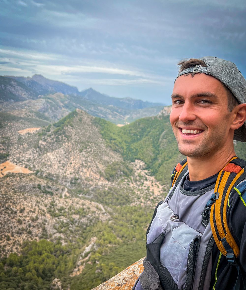 Jon Miksis smiling in a cap and hiking pack on the way up to Castell d'Alaró, with forested Tramuntana mountain ridges behind him